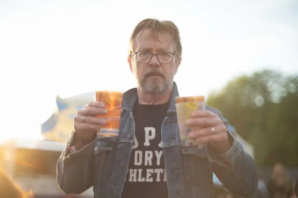 Ein Mann mit Jeansjacke hält zwei Becher Bier.